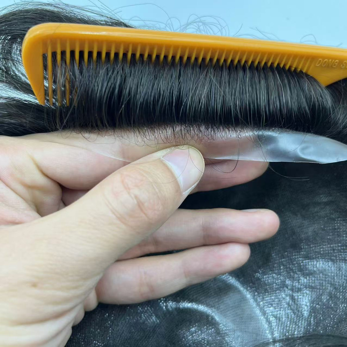 Side view of transparent 0.06-0.08mm thin poly skin base edge held beside knotted hair, showing base thickness